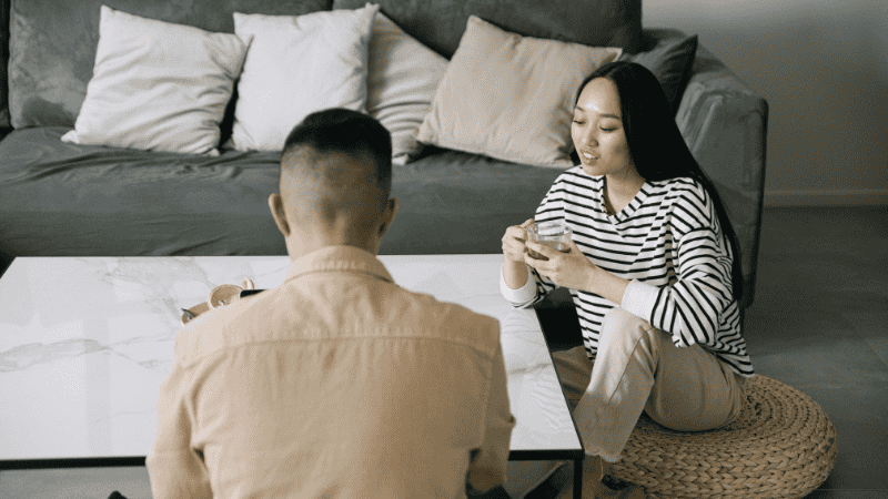A soft, neutral-toned illustration of two people sitting comfortably on a sofa engaged in an open, supportive conversation about sexual health and wellness.